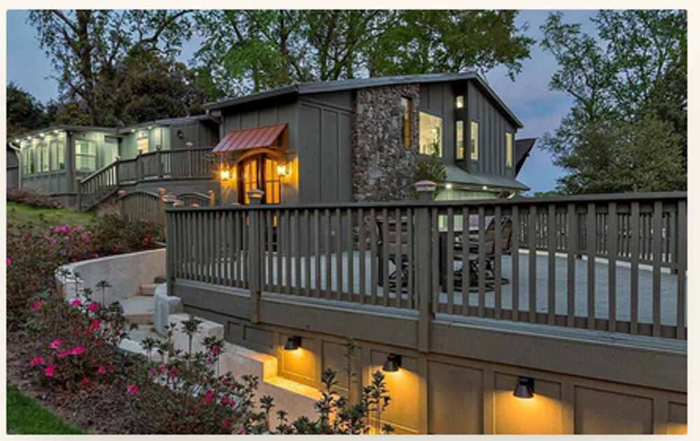 The Wilkshire House at twilight: warm interior windows glowing, copper-roofed arched entry lit, stone chimney illuminated, the wraparound deck set with chairs, and a soft line of landscape lights running along the base of the lower level and down the garden stairs, with pink azaleas in bloom in the foreground