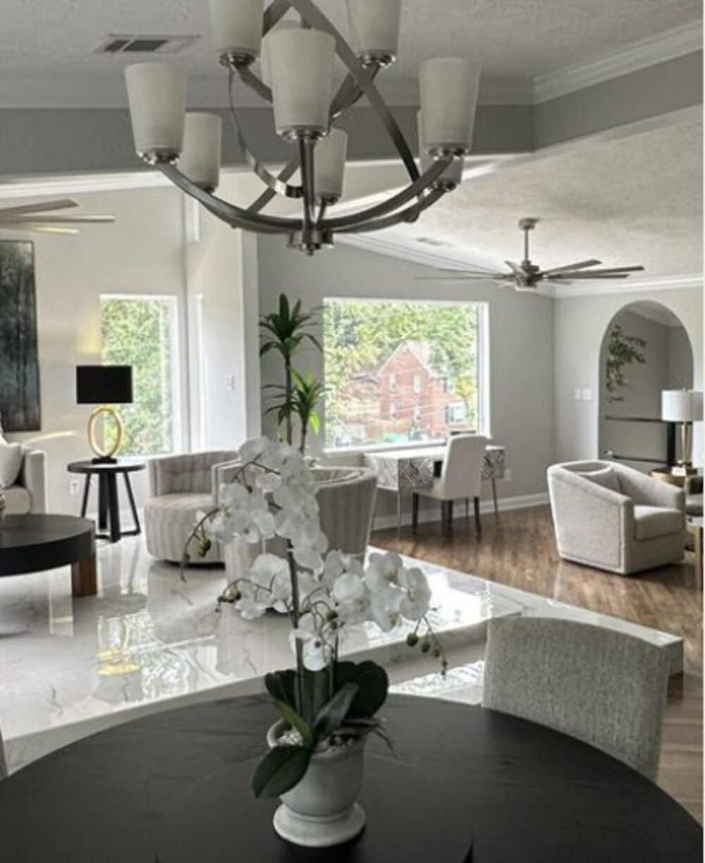 The main home's great room, viewed from the dining area through the arched entry — chandelier, ceiling fan, layered seating, and tree windows beyond